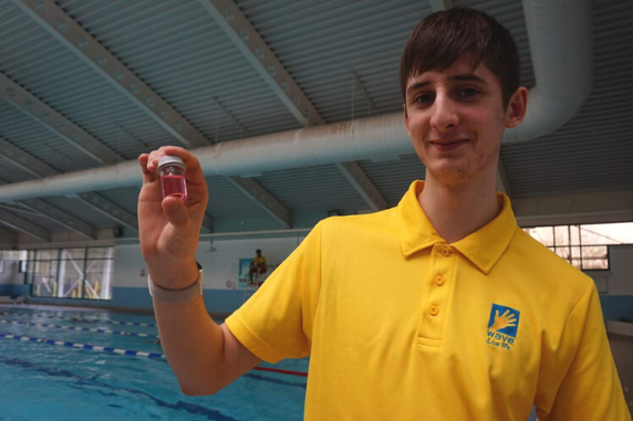 a young man in a lifeguard uniform next to a pool holding a small container of water